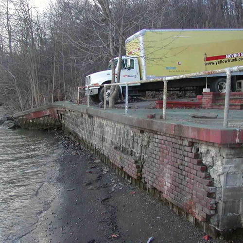 Esopus before. A picture of a waterfront property to be restored, with a crumbling brick wall against a river's edge, and a truck on top of the parking lot next to the wall. 
