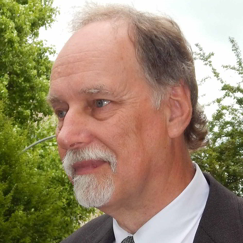 Headshot of Sven Hoeger. He has blue-grey eyes, greying-brown hair, a grey beard, and a slight tan. He is wearing a white shirt, tie, and dark jacket or sweater. It is a profile shot. There are trees and the sky behind his head. 